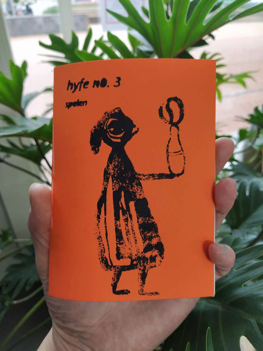 Foto van de kleine uitgave Hyfe nummer 3, met omslag van oranje karton en daarop in risoprint een figuur met wijde jurk of jas, die een arm uitsteekt met op hun hand een kegel waar een bal op balanceert, of misschien is het heel wat anders, je mag zelf weten hoe je het interpreteert. Ik, Eelkje houd de uitgave in mijn (witte) hand, op de achtergrond steekt het groen van een plant fel af tegen het oranje van het omslag.